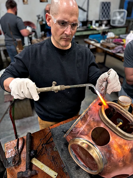 Nelson uses a torch to solder a part onto a copper helmet.