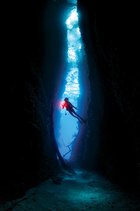 Diver explores a dark cavern. Their silhouette contrasts against the streak of sunlight.