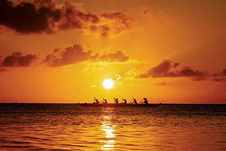 As the sun sinks toward the horizon, paddlers drive a traditional outrigger canoe.